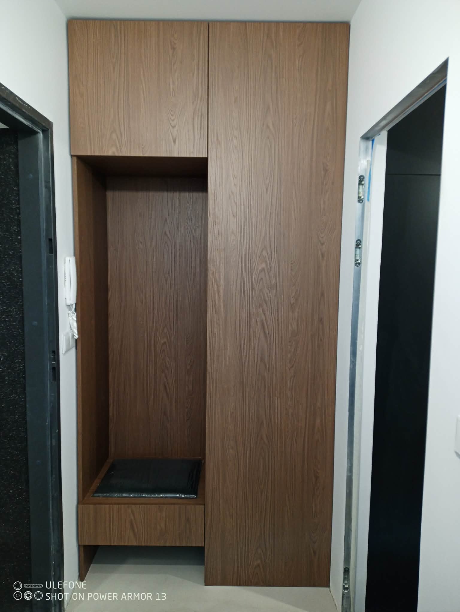 Funkcjonalna szafa wnękowa z siedziskiem w przedpokoju, wykończona okleiną drewnopodobną. Widoczny interkom i fragment ościeżnicy.