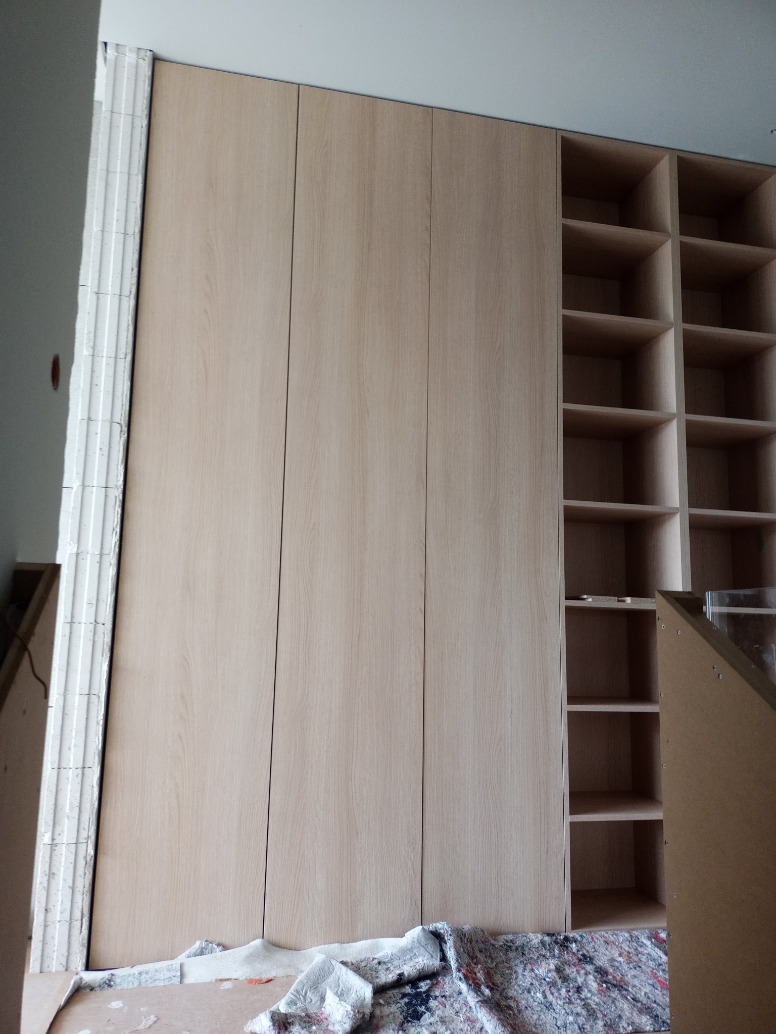 Jasna, drewniana szafa wnękowa z otwartymi półkami po prawej stronie, widoczna podczas montażu, z zabezpieczeniem podłogi. Minimalistyczny design.