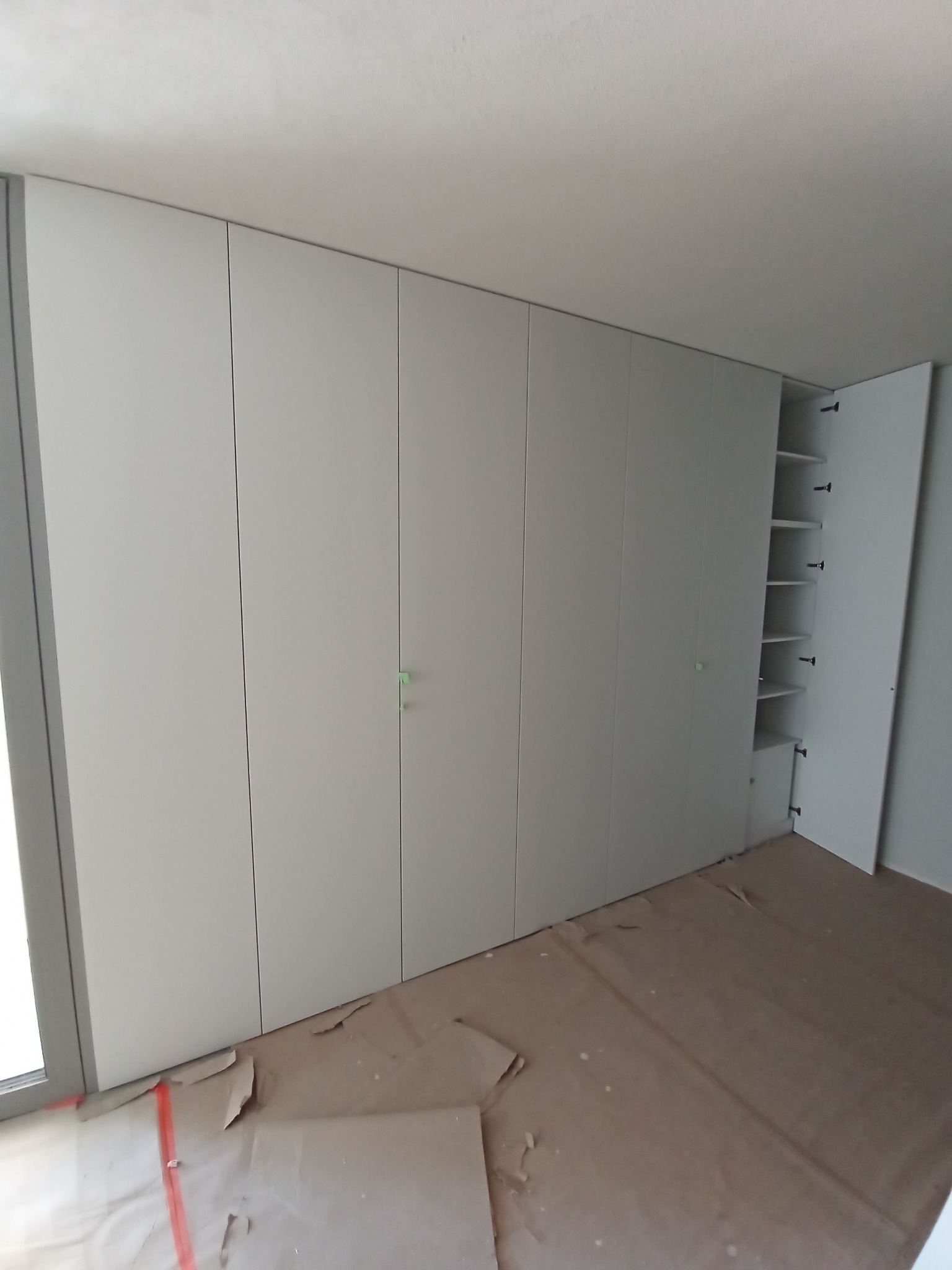 Długa, biała szafa wnękowa z otwartymi drzwiami ukazującymi półki. Podłoga zabezpieczona kartonem. Minimalistyczny design, jasne wnętrze.