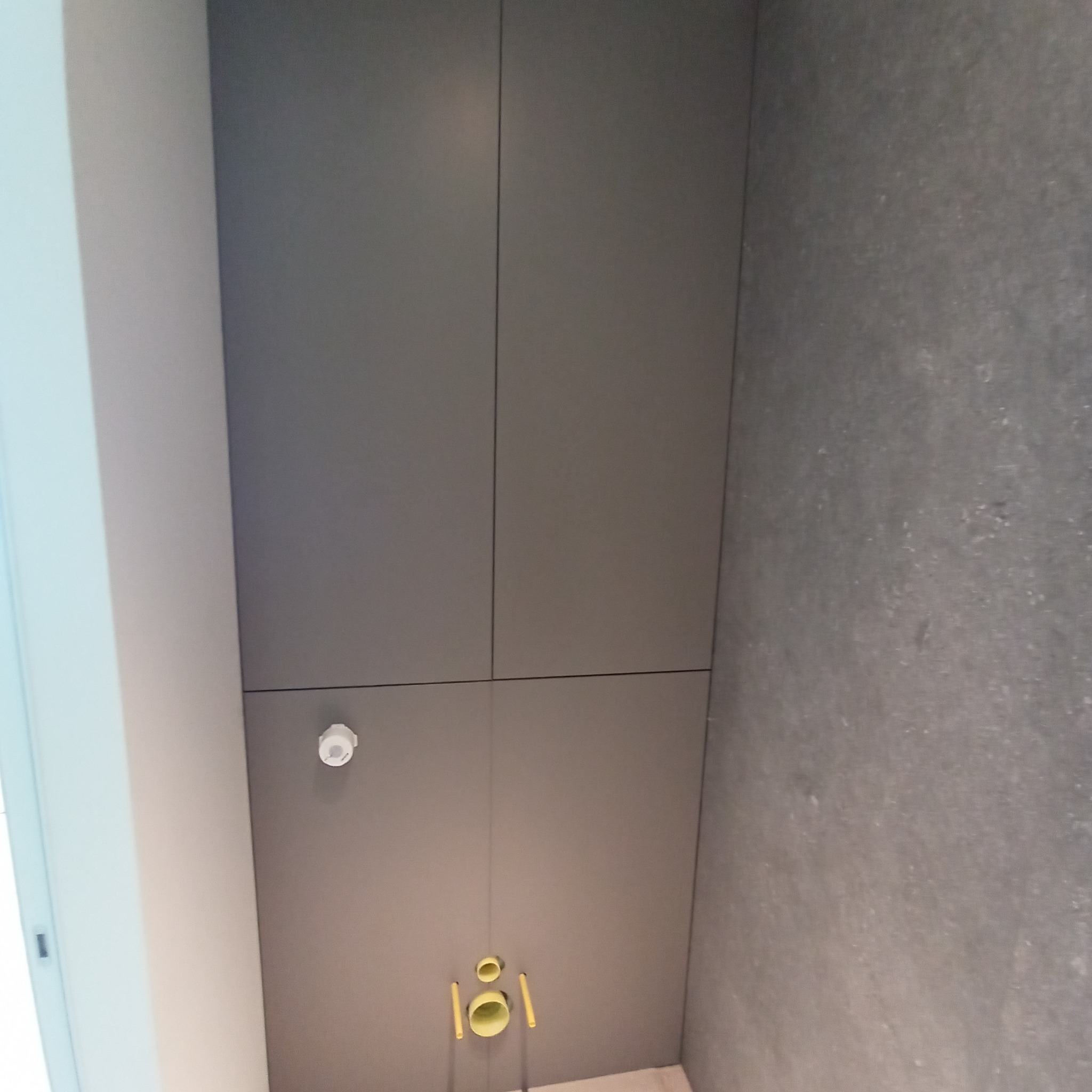 Szare panele ścienne z ukrytą instalacją sanitarną i czujnikiem dymu. Minimalistyczny design z widocznymi pionowymi liniami podziału paneli.