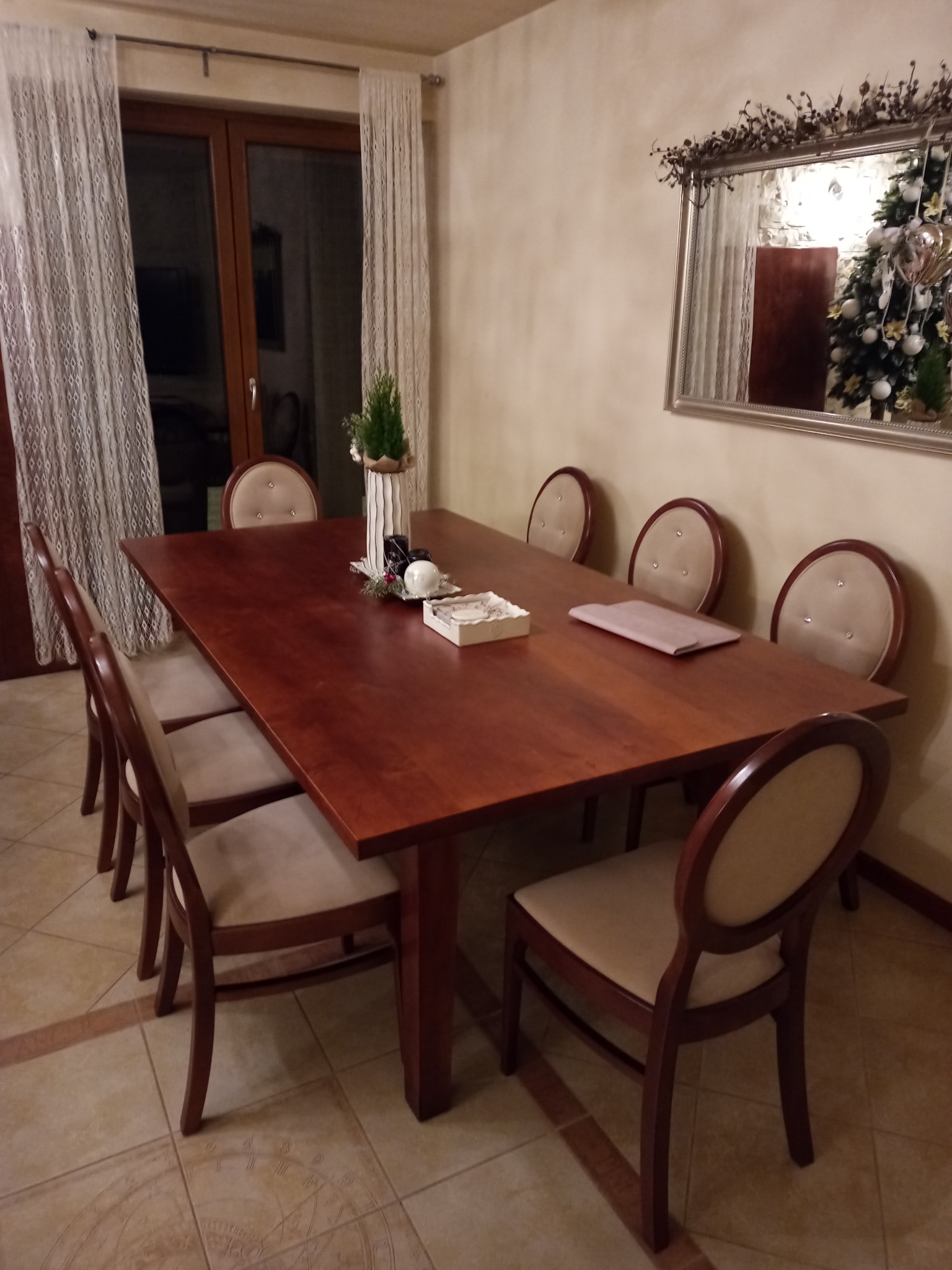 Drewniany stół z krzesłami w jadalni, nakryty do świątecznego posiłku. W tle okno z firankami i lustro z odbiciem choinki. Na podłodze wzór zegara słonecznego.