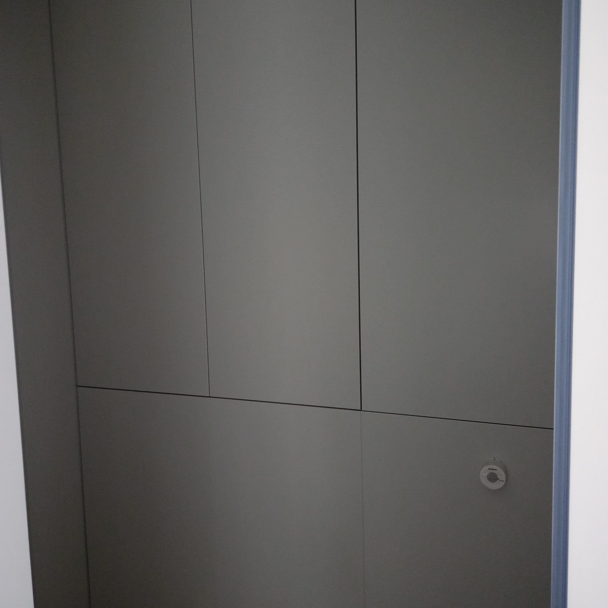 Szara, minimalistyczna szafa wnękowa z płaskimi frontami, z widocznym czujnikiem dymu. Proste linie i stonowana kolorystyka.