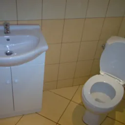 Biała umywalka z szafką i toaleta w łazience wyłożonej beżowymi płytkami.