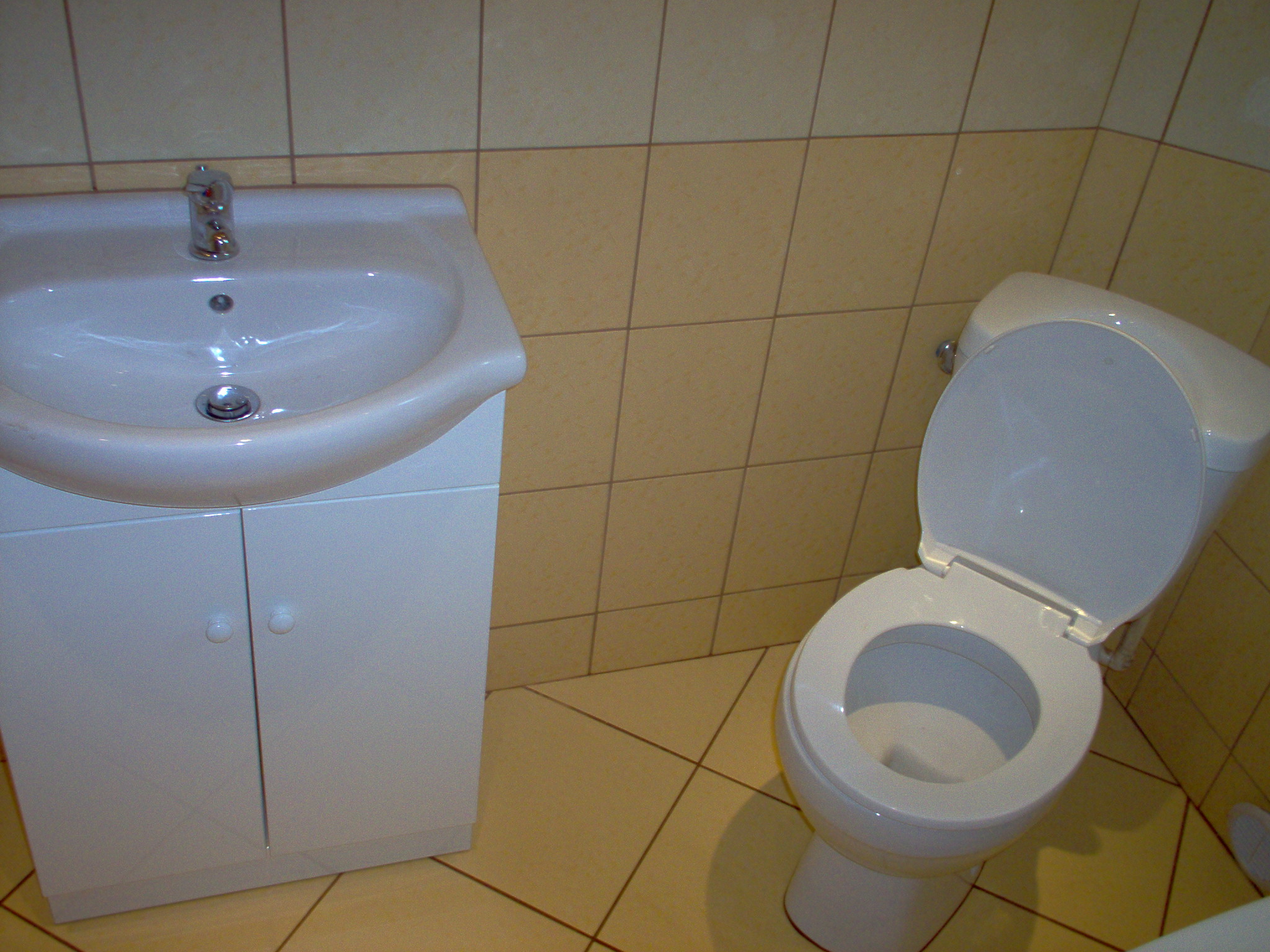 Biała umywalka z szafką i toaleta w łazience wyłożonej beżowymi płytkami.