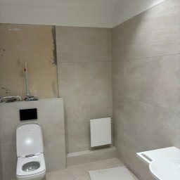 Andrzej Wiatr - Jasna łazienka w trakcie wykańczania: beżowe płytki imitujące kamień, podwieszana toaleta, grzejnik i umywalka. Część ściany bez płytek, widoczne narzędzia.