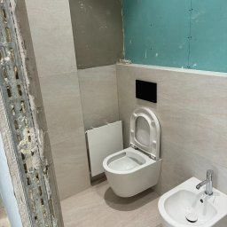 Andrzej Wiatr - Remont łazienki w trakcie: biała toaleta podtynkowa i bidet na tle beżowych płytek, widoczne płyty karton-gips i metalowy profil. Etap prac wykończeniowych.