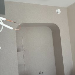 Andrzej Wiatr - Wnęka w ścianie z wyprowadzonymi instalacjami, widoczne przewody i rury. Ściana w jasnym odcieniu szarości, sufit z okrągłym oświetleniem. Przy podłodze widoczne torby i krzesło.