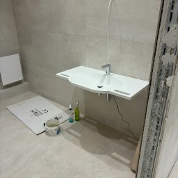 Andrzej Wiatr - Montaż umywalki w łazience z beżowymi płytkami. Widoczne narzędzia i materiały budowlane. Umywalka zamontowana, ale prace wykończeniowe trwają.