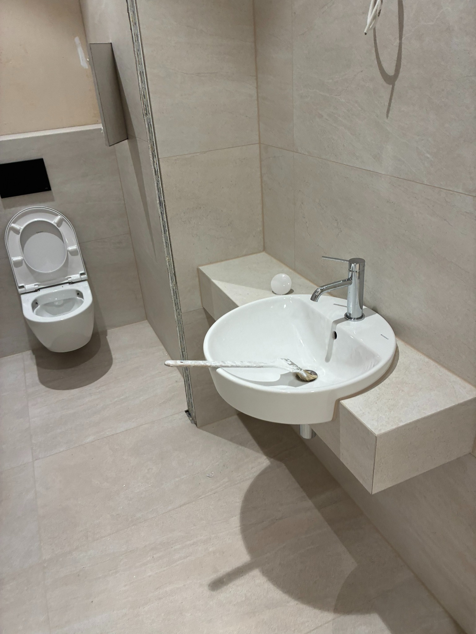 Jasna łazienka po remoncie: podwieszana toaleta, umywalka z baterią na blacie z płytek, płytki na ścianach i podłodze. Widoczny pędzel w umywalce.