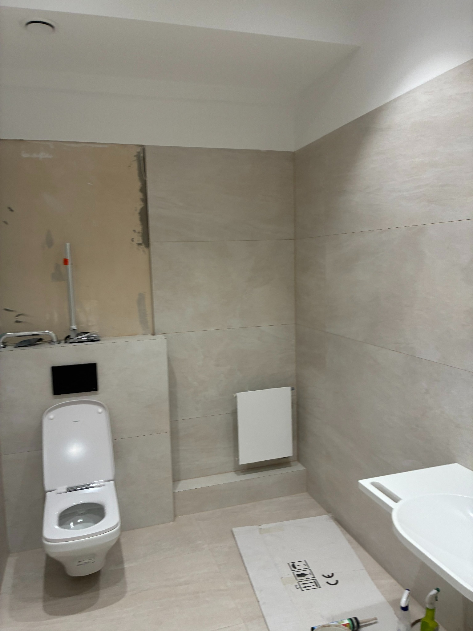 Jasna łazienka w trakcie wykańczania: beżowe płytki imitujące kamień, podwieszana toaleta, grzejnik i umywalka. Część ściany bez płytek, widoczne narzędzia.