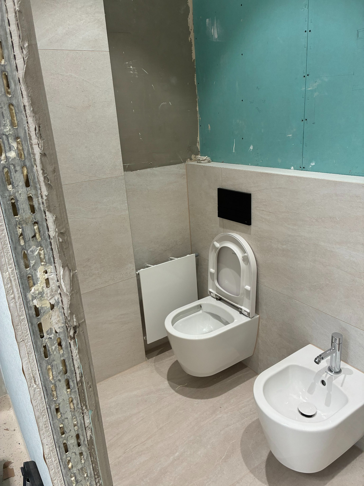 Remont łazienki w trakcie: biała toaleta podtynkowa i bidet na tle beżowych płytek, widoczne płyty karton-gips i metalowy profil. Etap prac wykończeniowych.