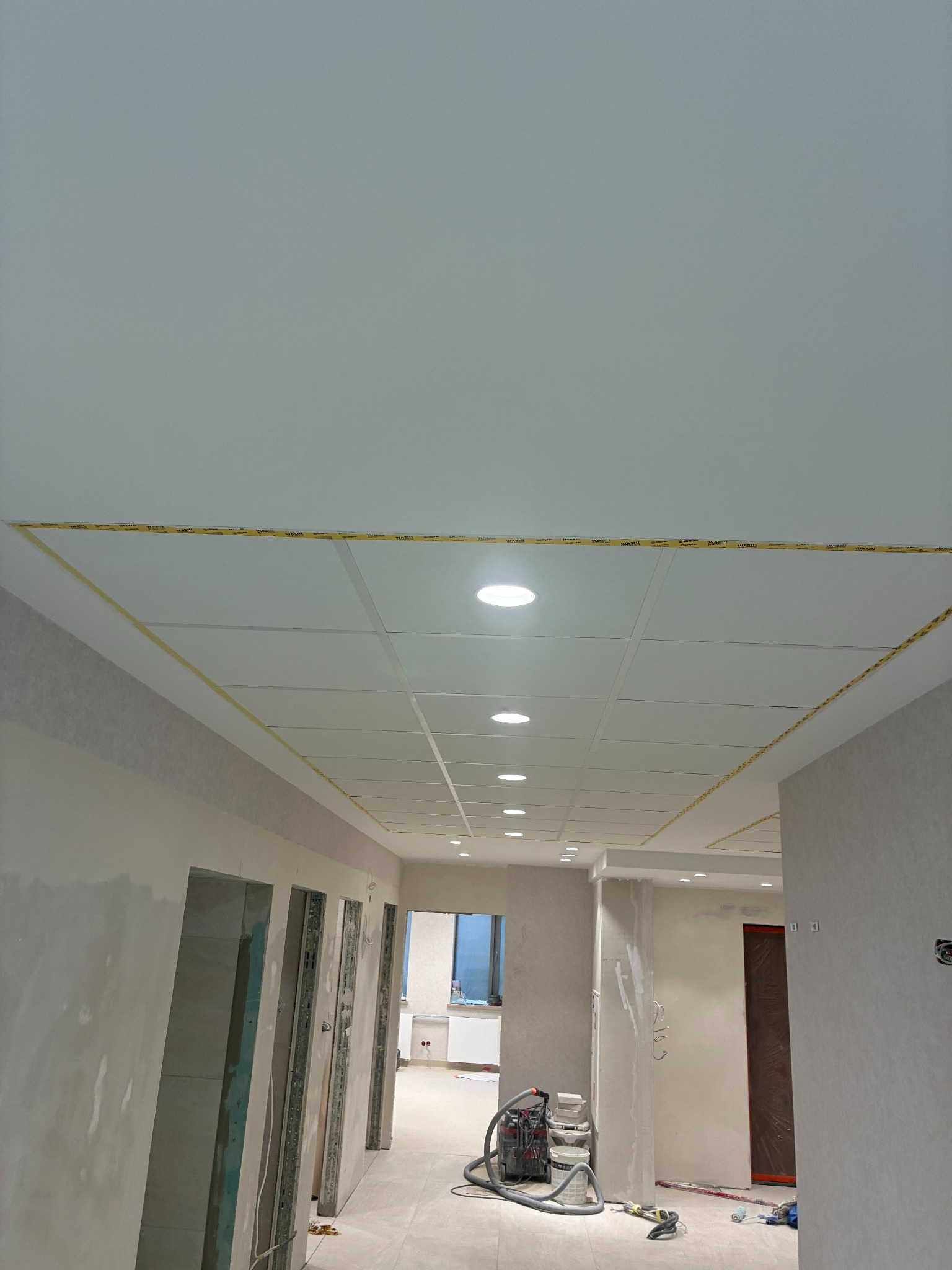 Sufit podwieszany z oświetleniem punktowym w trakcie montażu, zabezpieczony taśmą. Ściany przygotowane do malowania, widoczny sprzęt budowlany.