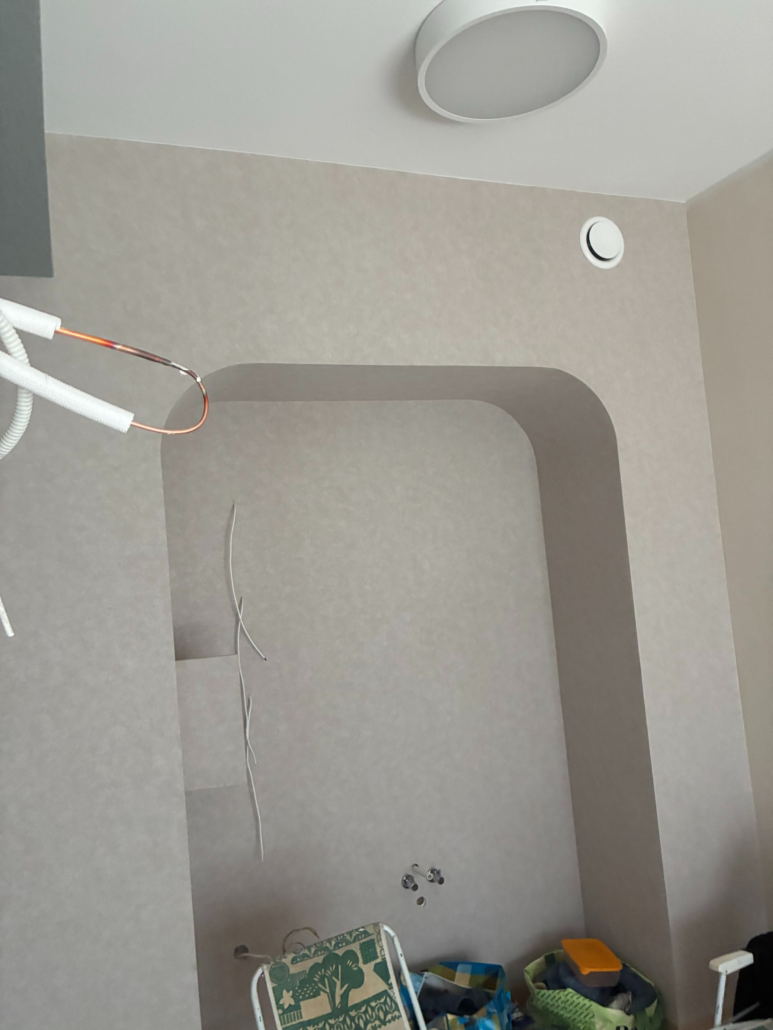 Wnęka w ścianie z wyprowadzonymi instalacjami, widoczne przewody i rury. Ściana w jasnym odcieniu szarości, sufit z okrągłym oświetleniem. Przy podłodze widoczne torby i krzesło.