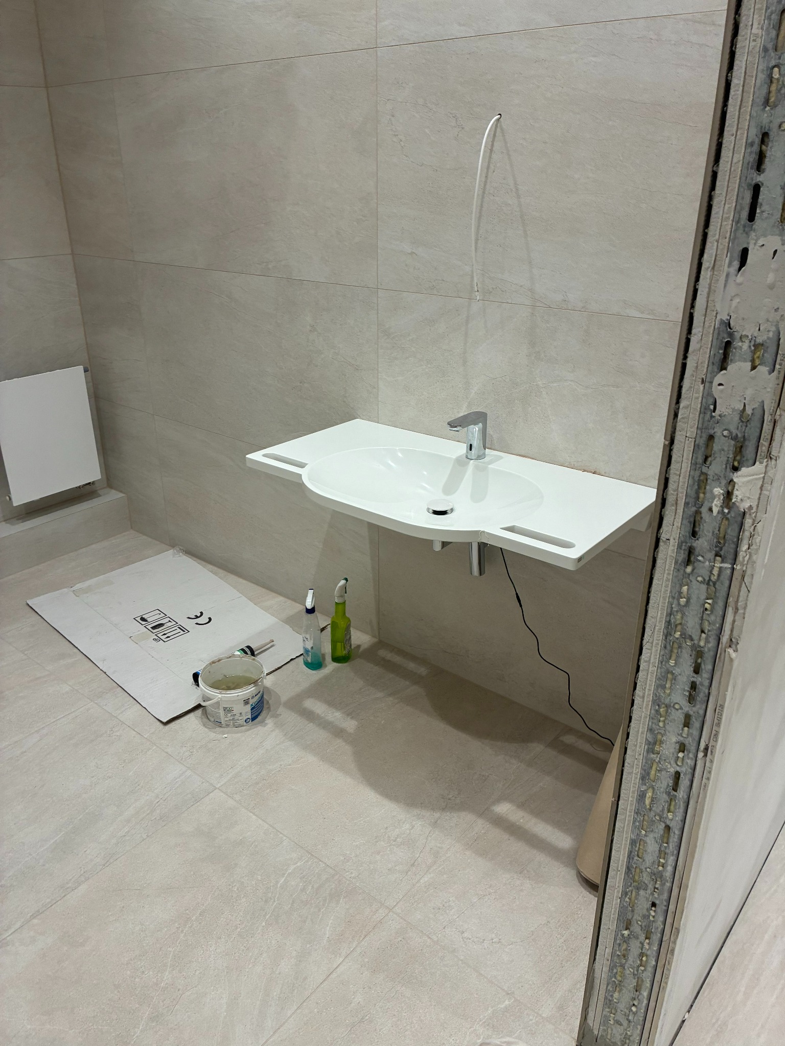 Montaż umywalki w łazience z beżowymi płytkami. Widoczne narzędzia i materiały budowlane. Umywalka zamontowana, ale prace wykończeniowe trwają.