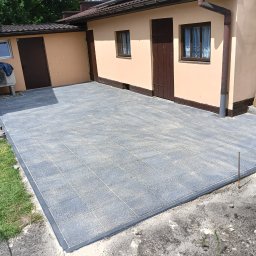 GREENGARDEN Łukasz Jaskólski - Ułożona szara kostka brukowa na podwórku przy budynku z brązowymi drzwiami i oknami. Widoczna trawa, narzędzia i elementy wykończeniowe.