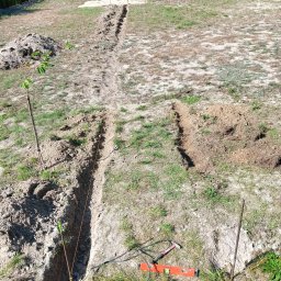 GREENGARDEN Łukasz Jaskólski - Wykopane rowy w ziemi, prawdopodobnie pod instalację nawadniającą. Widoczne narzędzia: poziomica, młotek, sznurek wyznaczający linię. W tle drzewa i ogrodzenie.