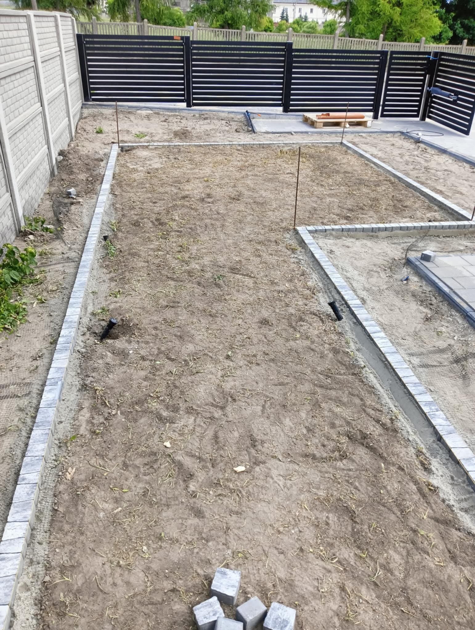 Przygotowanie terenu pod kostkę brukową: obrzeża ułożone, teren wyrównany, widoczne rury nawadniające. W tle nowoczesne ogrodzenie panelowe i dom.