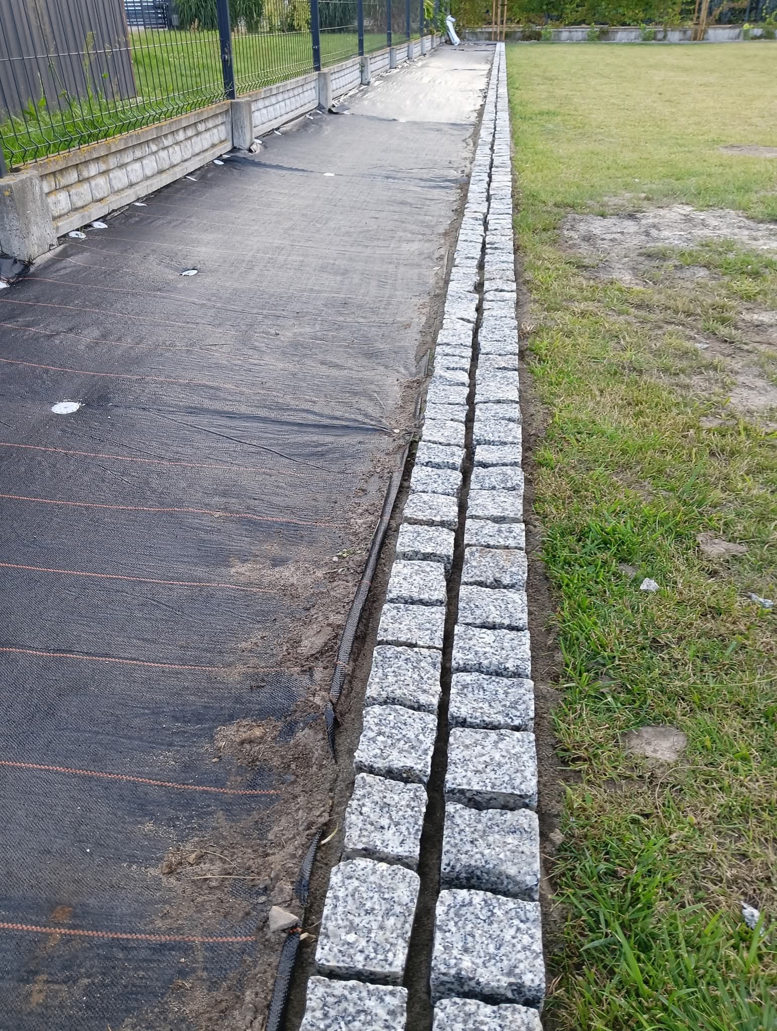 Granitowa obwódka z kostki, oddzielająca trawnik od agrowłókniny, widok z góry, lekki kąt. Element wykończeniowy ogrodu, surowy kamień.