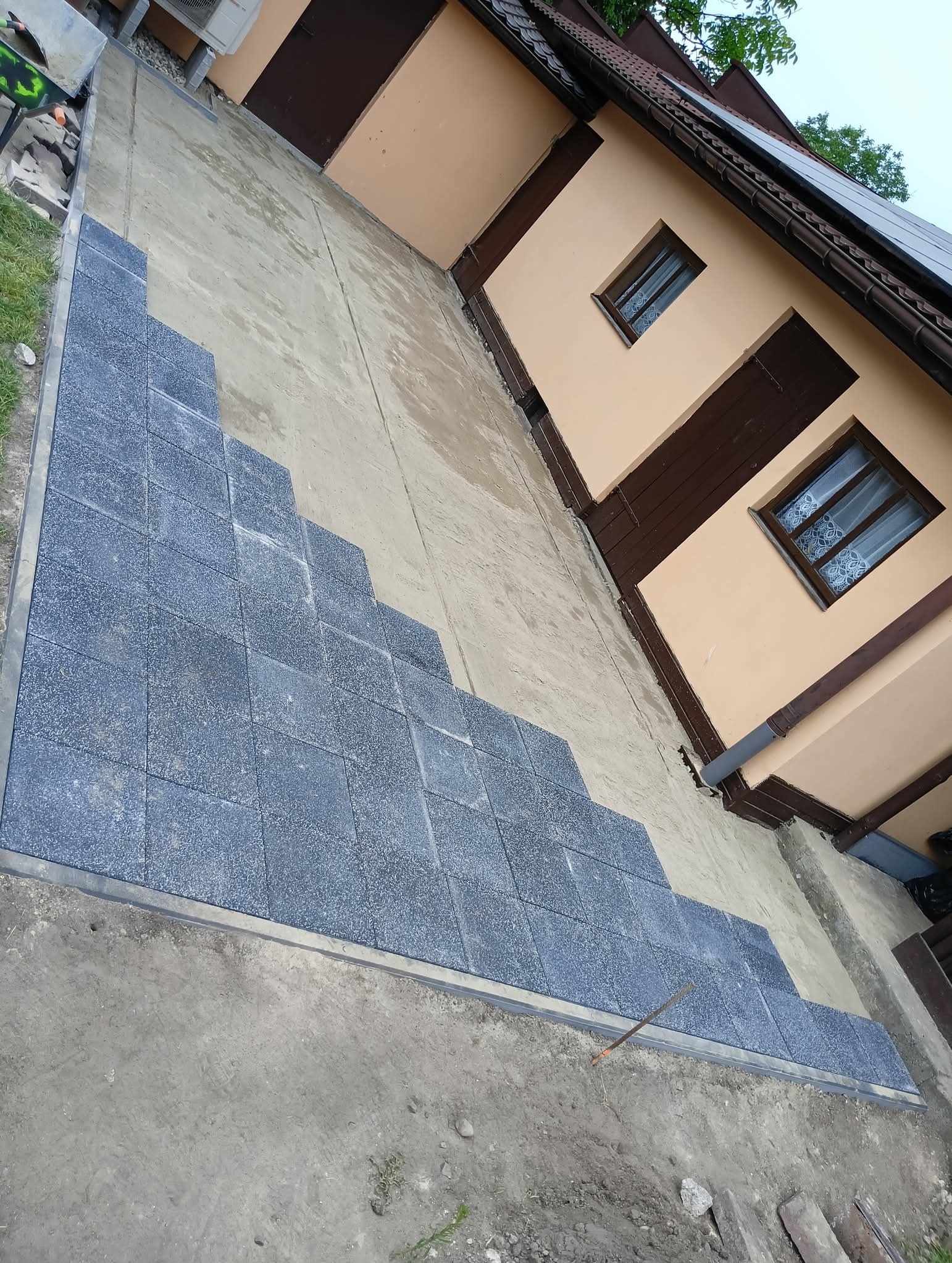 Częściowo ułożona, szara kostka brukowa na podwórku przy domu. Widoczne krawężniki i przygotowane podłoże. Ujęcie z góry, ukośne.