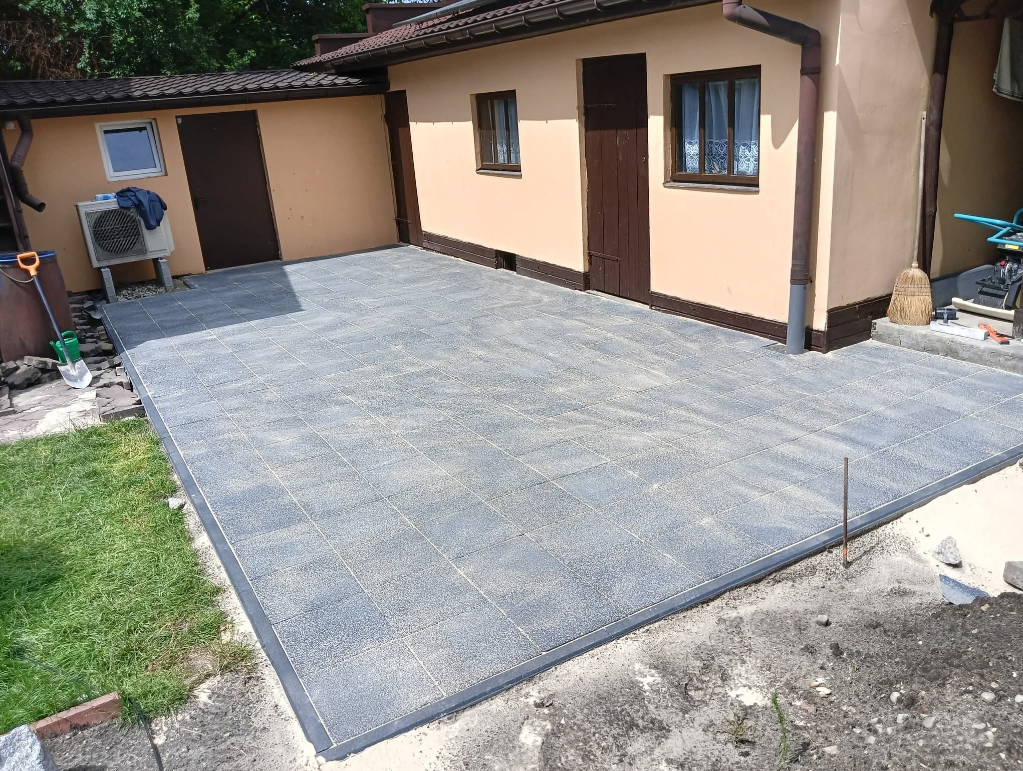 Ułożona szara kostka brukowa na podwórku przy budynku z brązowymi drzwiami i oknami. Widoczna trawa, narzędzia i elementy wykończeniowe.