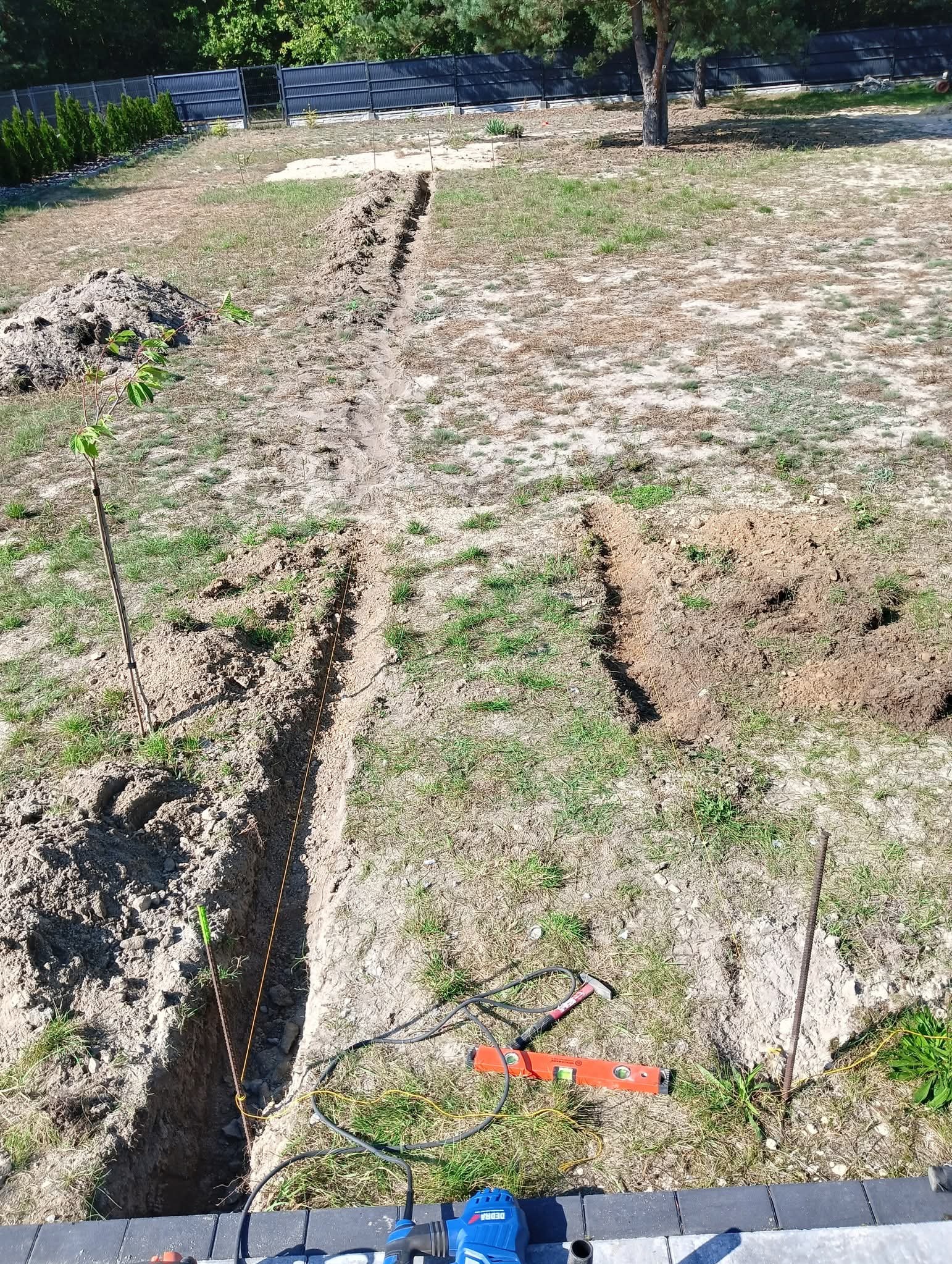 Wykopane rowy w ziemi, prawdopodobnie pod instalację nawadniającą. Widoczne narzędzia: poziomica, młotek, sznurek wyznaczający linię. W tle drzewa i ogrodzenie.