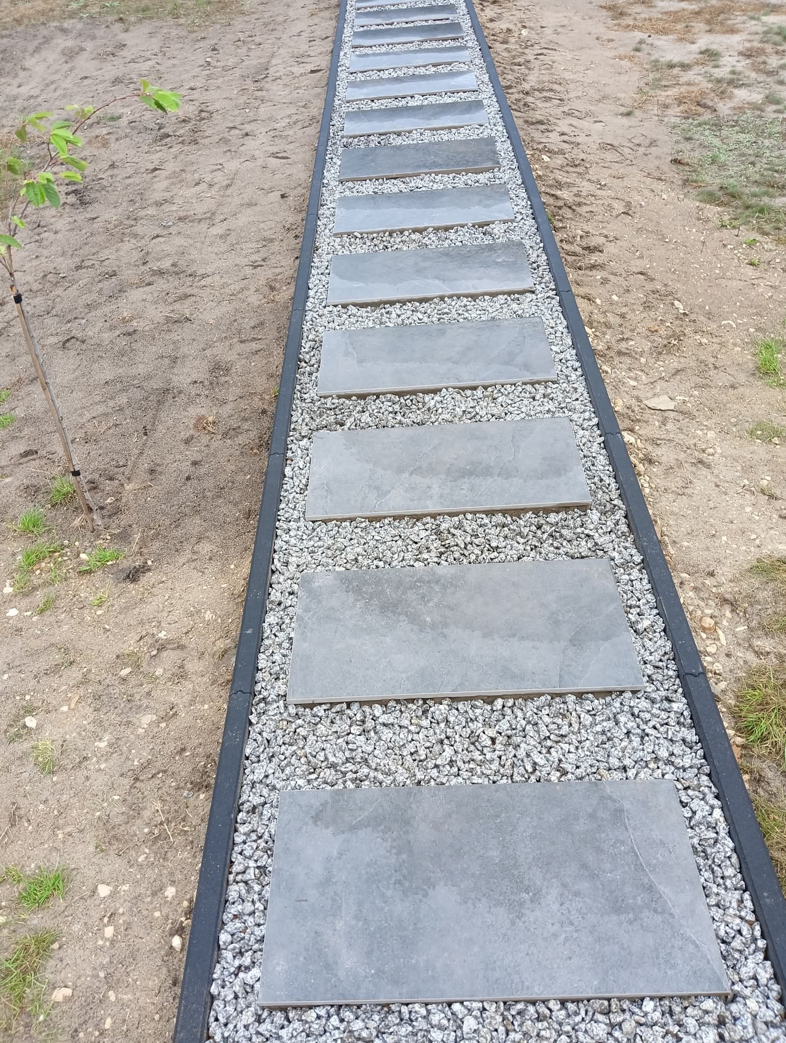 Ścieżka z szarych płyt ułożonych na drobnym żwirze, obramowana czarnym obrzeżem. W tle ziemia i małe drzewko. Widok z góry, perspektywa zbieżna.