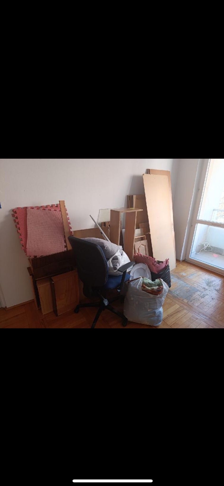 Pokój przed opróżnianiem: zagracony fotel biurowy, worki z rzeczami, meble i panele pod ścianą, parkiet z widocznymi śladami użytkowania, okno balkonowe.