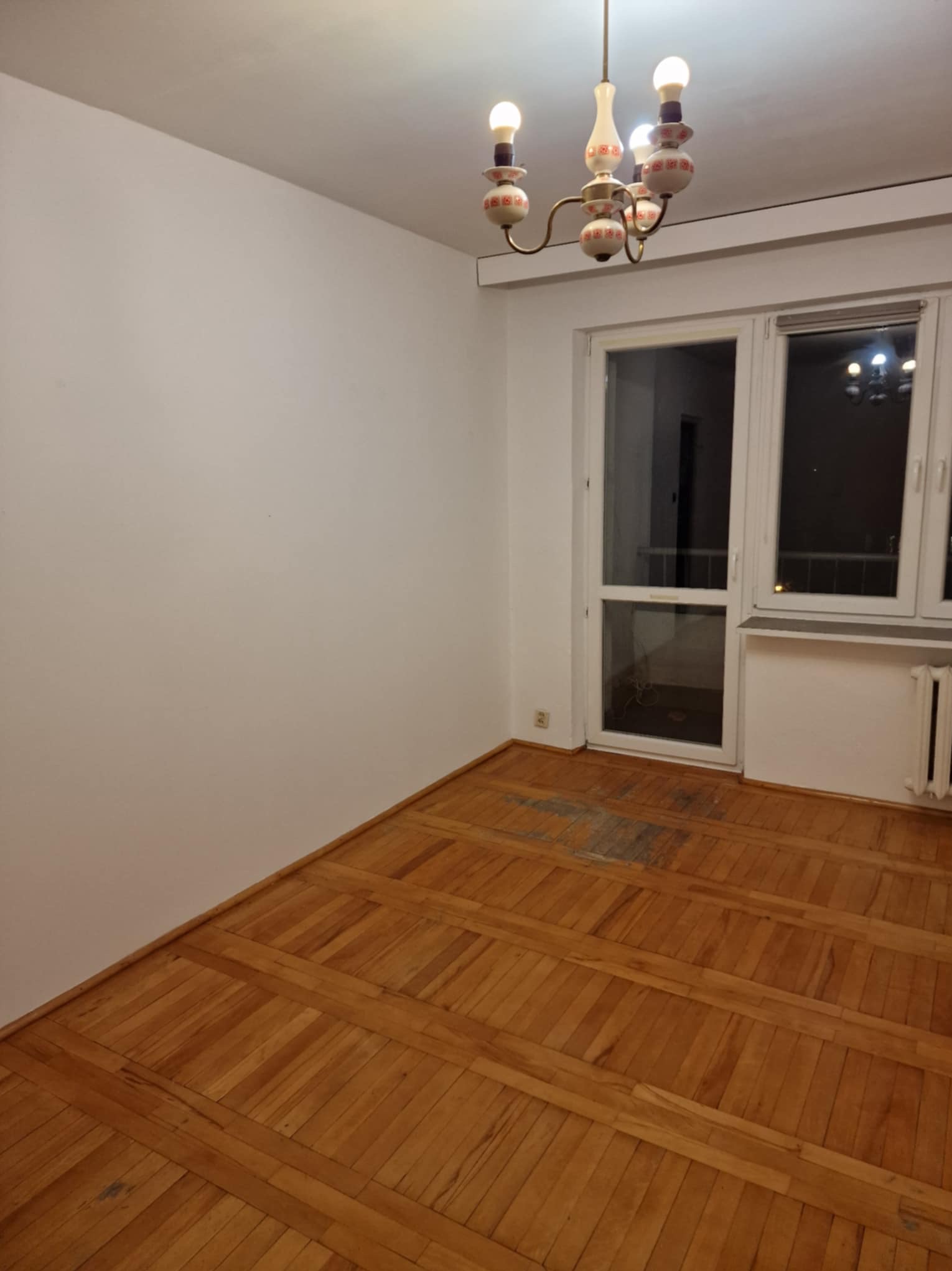 Pusty pokój z parkietem i białymi ścianami. Widoczny żyrandol w stylu retro, okno z grzejnikiem. Podłoga z widocznymi śladami użytkowania.