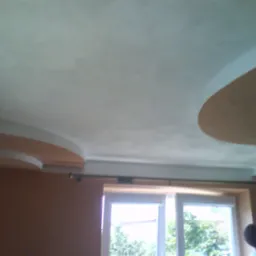 Wykończony sufit z elementami dekoracyjnymi z płyty karton gips w jasnych kolorach, widoczny fragment okna i sylwetka osoby w tle.