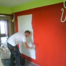 Mężczyzna w białej koszulce maluje czerwoną ścianę na biało, widoczna drabina, puszki z farbą i narzędzia malarskie, wnętrze w trakcie odświeżania.