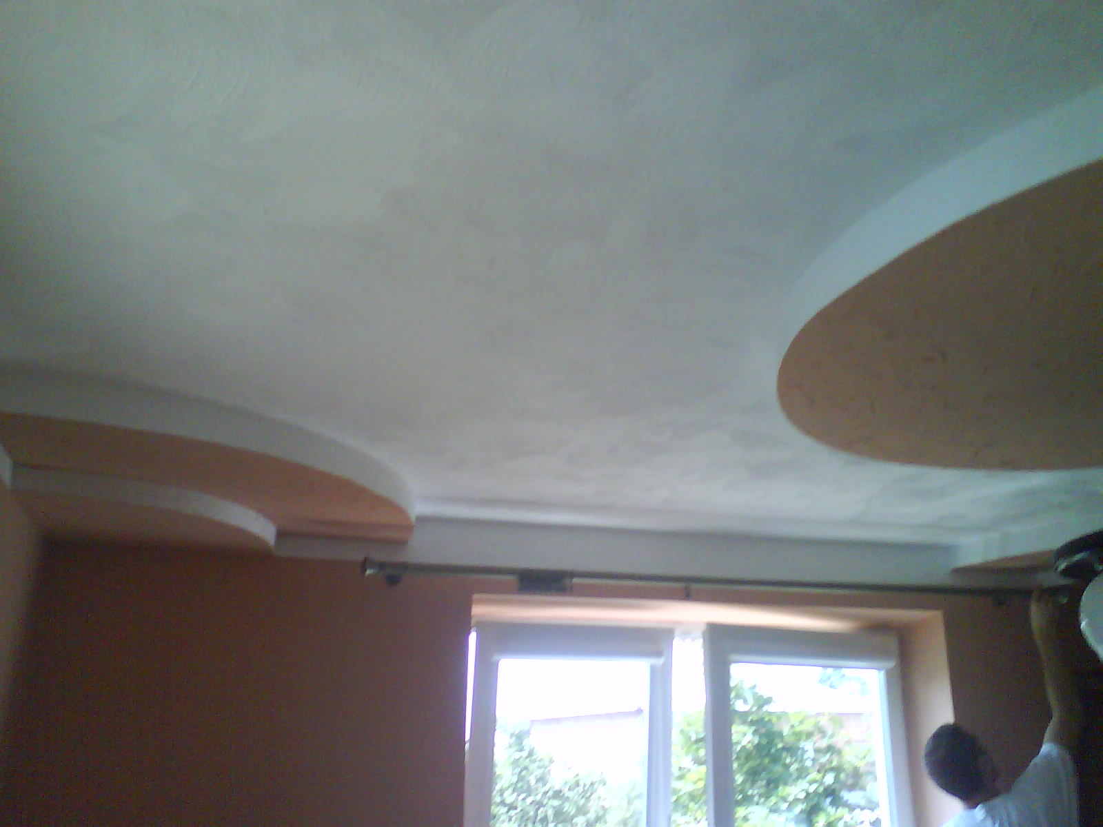 Wykończony sufit z elementami dekoracyjnymi z płyty karton gips w jasnych kolorach, widoczny fragment okna i sylwetka osoby w tle.