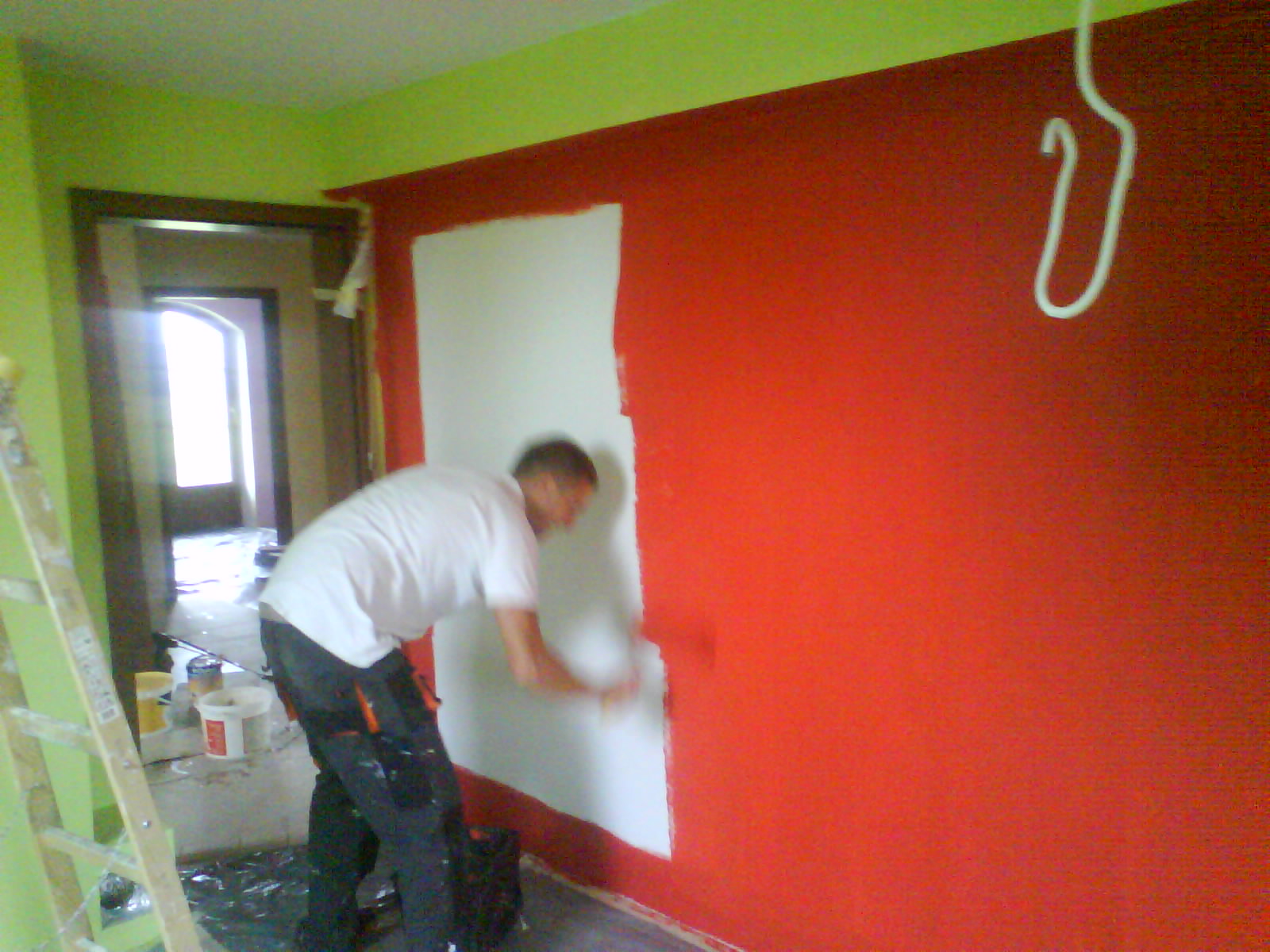 Mężczyzna w białej koszulce maluje czerwoną ścianę na biało, widoczna drabina, puszki z farbą i narzędzia malarskie, wnętrze w trakcie odświeżania.