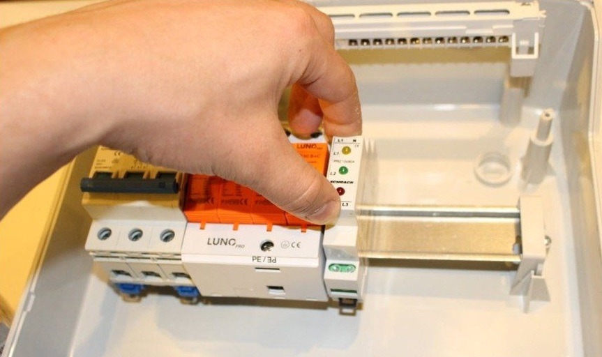 Montaż ogranicznika przepięć LUNC PRO w skrzynce elektrycznej. Widoczna dłoń instalatora i szyna montażowa. Elementy instalacji w kolorze pomarańczowym i białym.