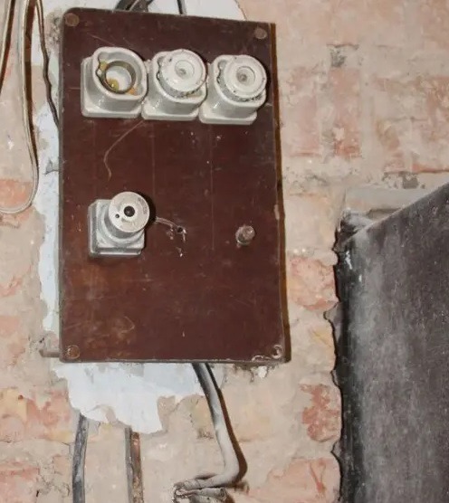 Stara, brązowa tablica z ceramicznymi bezpiecznikami na tle ceglanej ściany. Widoczne przewody elektryczne i ślady napraw. Jeden bezpiecznik jest uszkodzony.