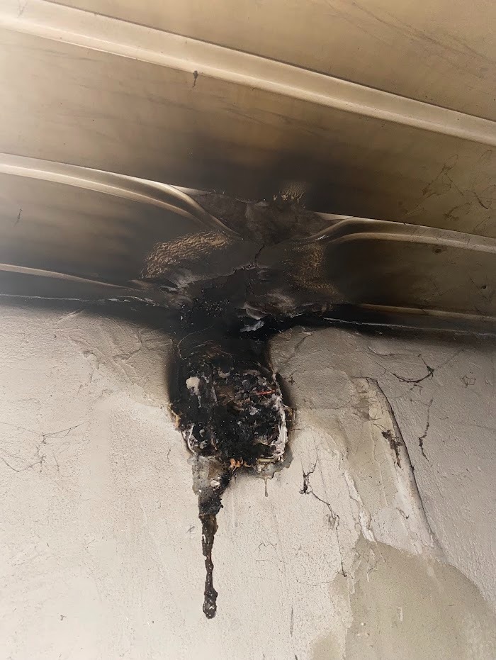 Przepalona instalacja elektryczna z widocznymi śladami pożaru na ścianie i suficie. Uszkodzone przewody i stopiony plastik. Niebezpieczeństwo zwarcia i porażenia.