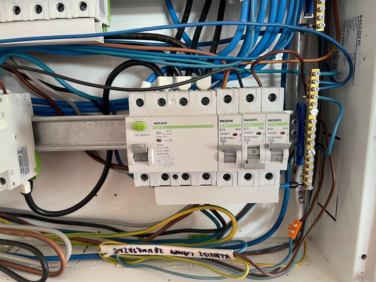 Rozdzielnica elektryczna z bezpiecznikami Noark, okablowaniem w kolorach niebieskim, brązowym i żółto-zielonym. Widoczne oznaczenia i parametry techniczne.