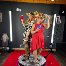 Robert Osóbka - Dwie kobiety w kostiumach pozują na platformie fotobudki 360 z rekwizytami: pistoletem na bańki i atrapami pieniędzy. Czerwony dywan i oświetlenie LED.