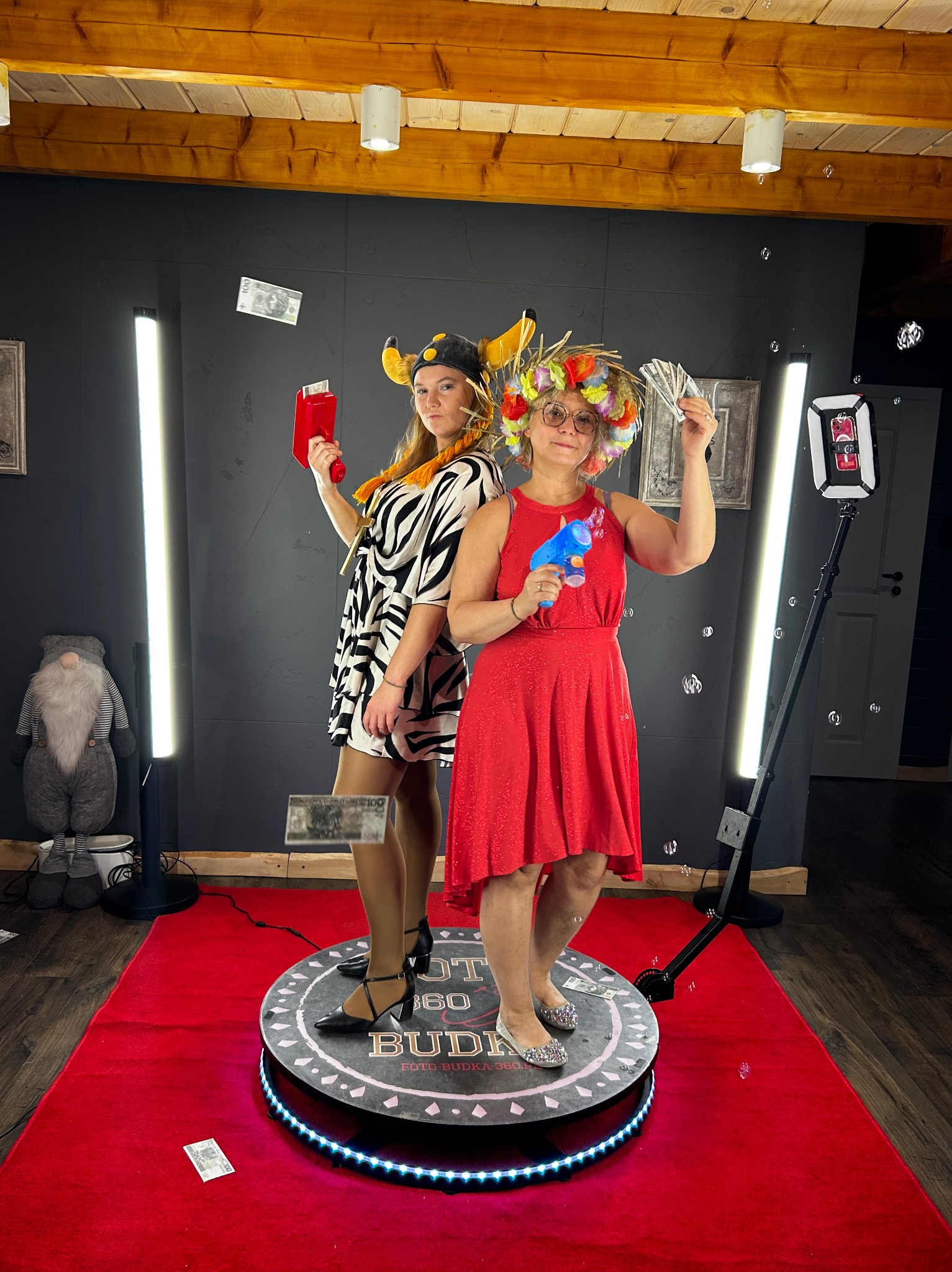 Dwie kobiety w kostiumach pozują na platformie fotobudki 360 z rekwizytami: pistoletem na bańki i atrapami pieniędzy. Czerwony dywan i oświetlenie LED.