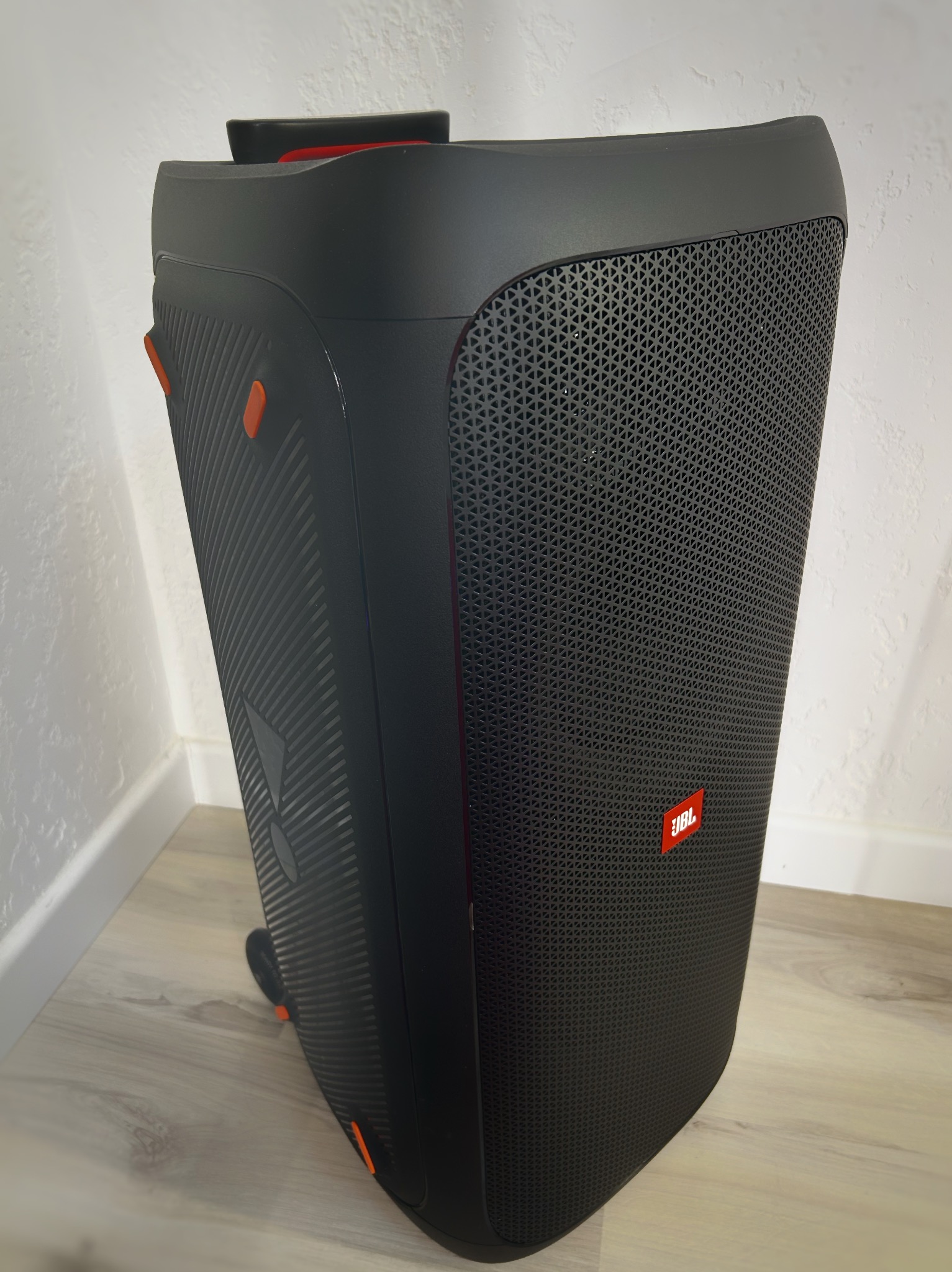 Czarny głośnik JBL Partybox 310 z wysuwaną rączką, ustawiony na podłodze, widoczny z boku, z logo JBL w prawym dolnym rogu. Tło stanowi biała ściana.