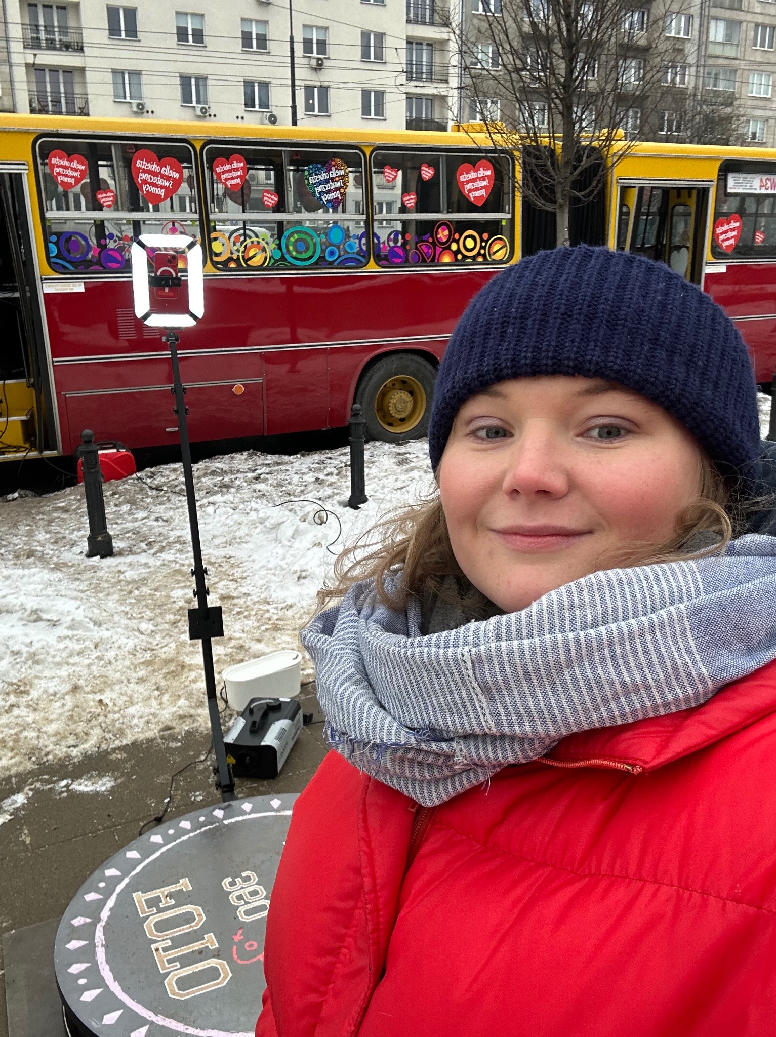 Uśmiechnięta kobieta w zimowej czapce na tle czerwonego autobusu z dekoracjami i sprzętu fotobudki 360, w zimowej scenerii.