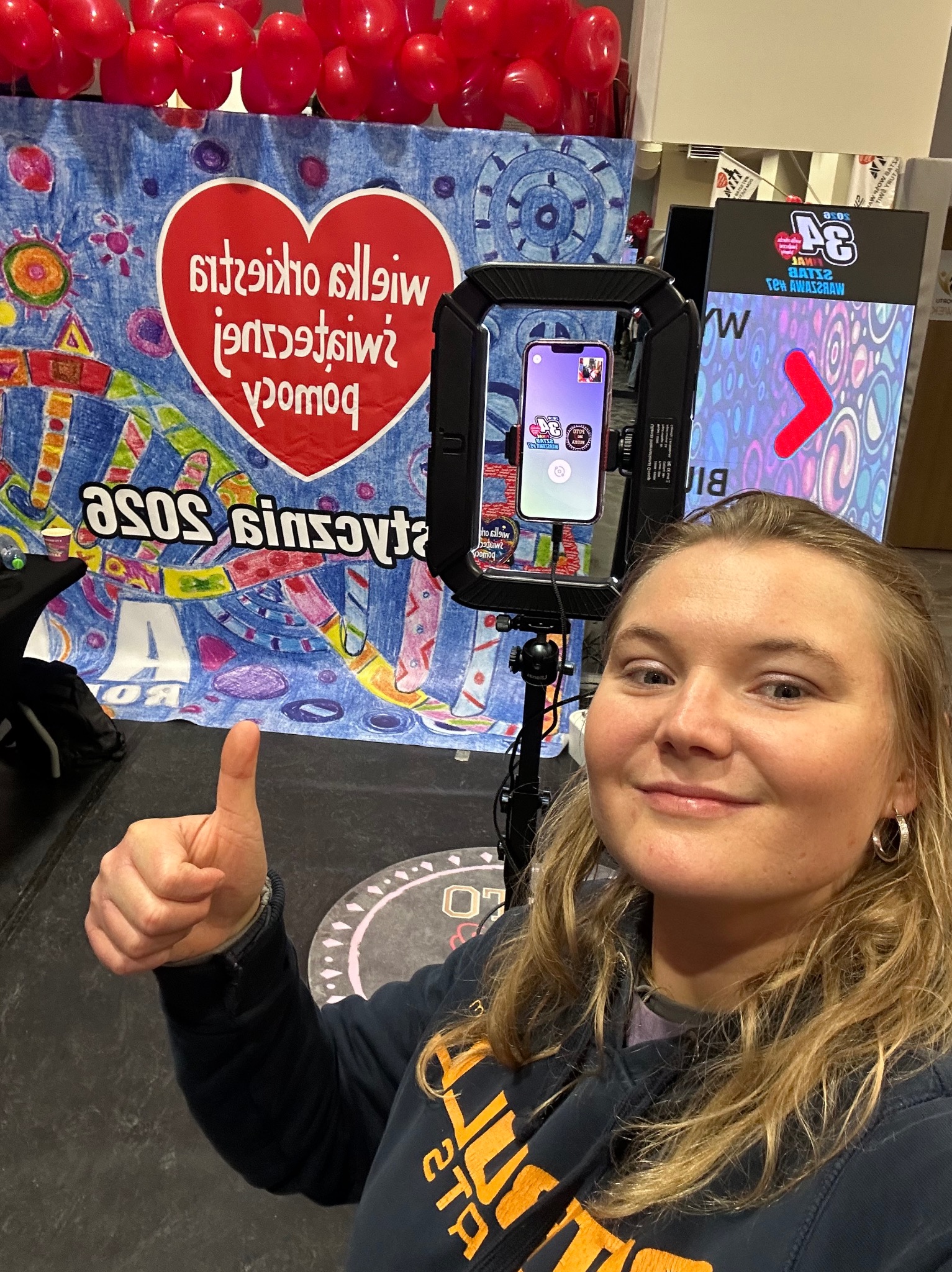 Uśmiechnięta kobieta robi selfie z fotobudką WOŚP. W tle kolorowy baner i balony. Kciuk w górę. Telefon w uchwycie z Hello Kitty. Dobra zabawa na imprezie.