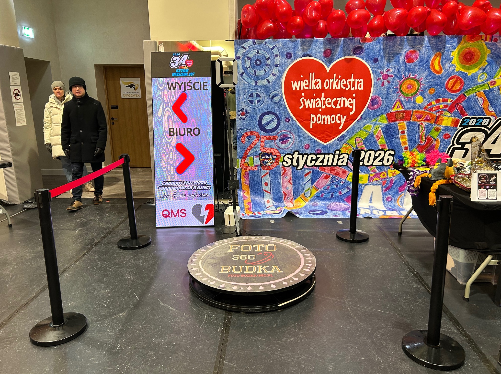 Okrągła fotobudka 360 na tle scenografii WOŚP z balonami i plakatem. Widoczni ludzie w tle i elektroniczny szyld z napisem 'Wyjście' i 'Biuro'.