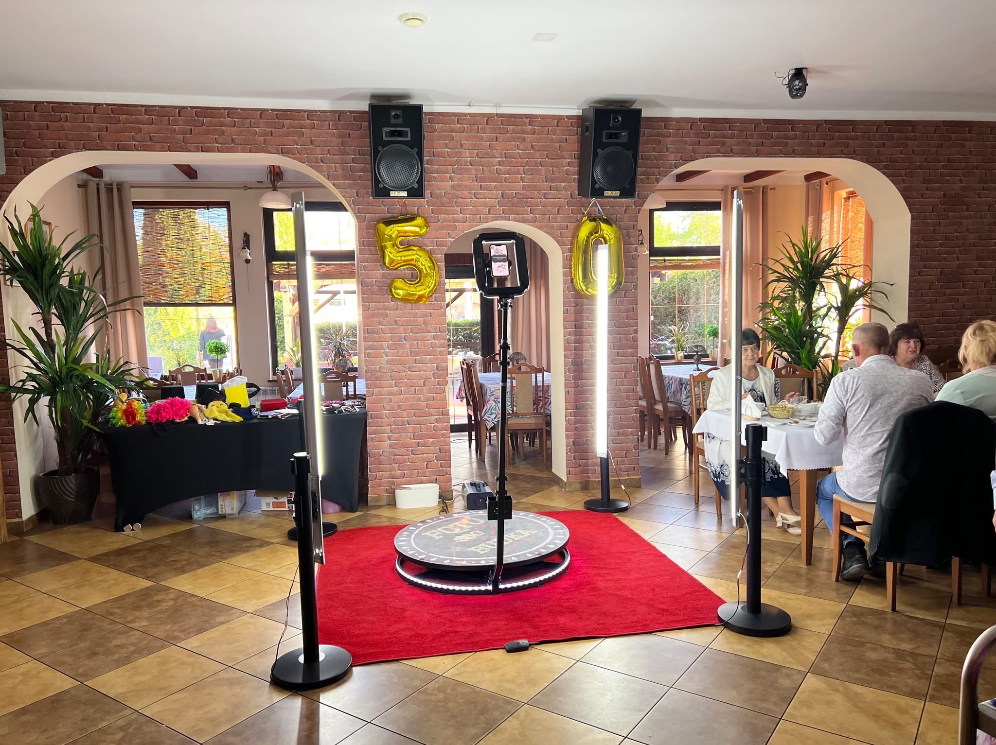 Sala z fotobudką 360 na czerwonym dywanie, dekoracje urodzinowe (balony cyfry 50), goście przy stole. Fotobudka z oświetleniem LED i uchwytem na telefon.