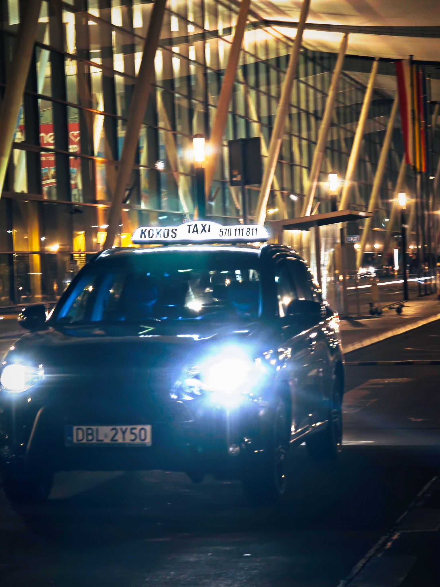 Nocne ujęcie taksówki Kokos Taxi z Bolesławca przed nowoczesnym budynkiem z przeszkloną fasadą. Widoczne światła samochodu i tablica z numerem telefonu.