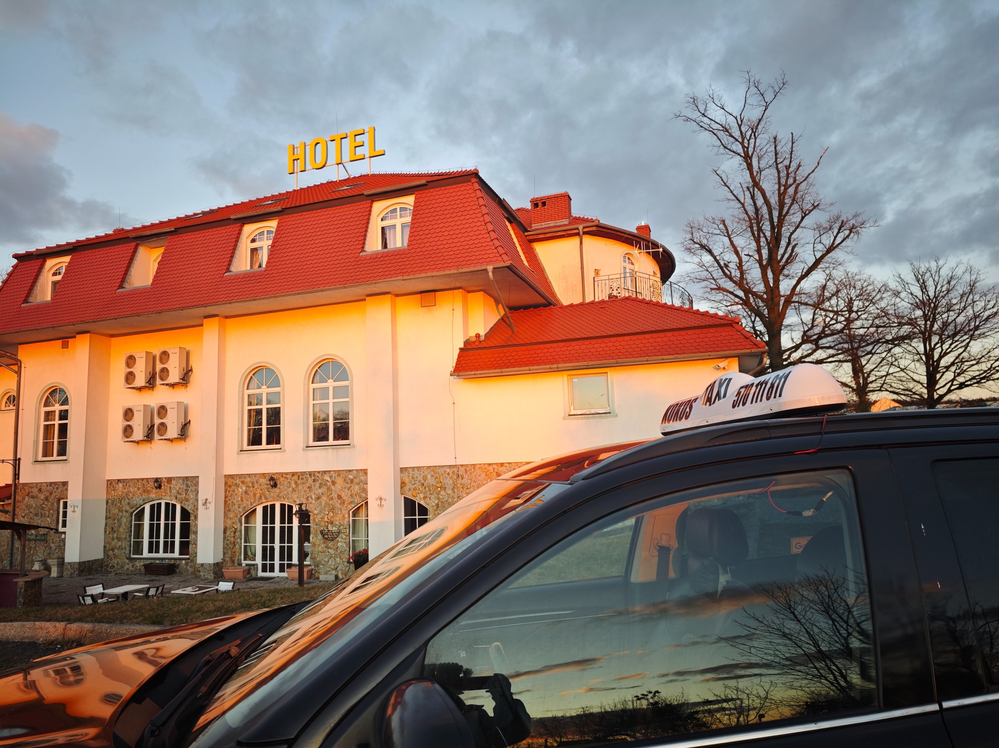 Czarny samochód taxi z napisem na dachu na tle budynku hotelu z czerwonym dachem i żółtym napisem 'HOTEL' o zmierzchu.