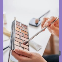 Wiktoria Kawa - Paleta cieni do powiek w dłoni, pędzel nabiera cień. Widoczne inne akcesoria do makijażu na białym stole. Delikatne, naturalne światło.