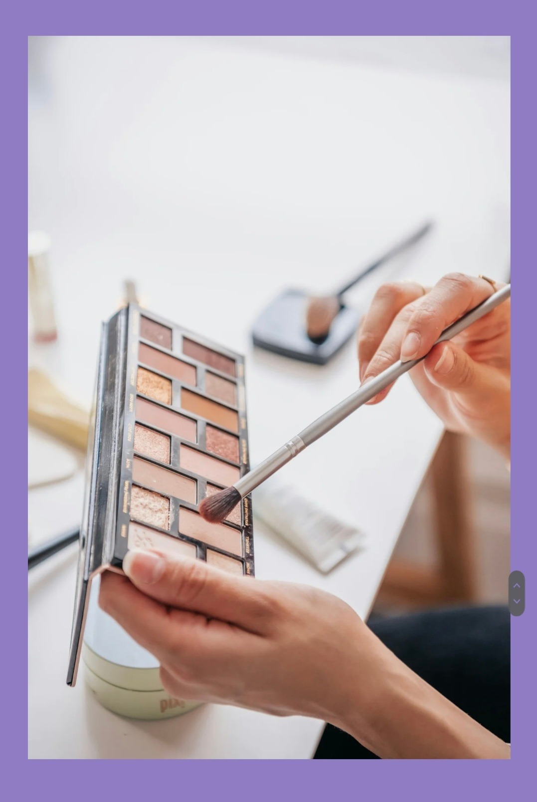 Paleta cieni do powiek w dłoni, pędzel nabiera cień. Widoczne inne akcesoria do makijażu na białym stole. Delikatne, naturalne światło.