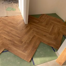 Mistrz-Bud - Układanie parkietu w jodełkę na zielonym podkładzie. Widoczny fragment ułożonej podłogi i przygotowane panele. Częściowo zabezpieczona ściana folią malarską.