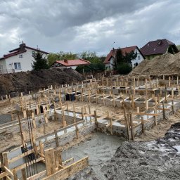 Mistrz-Bud - Wylane fundamenty pod budowę domu jednorodzinnego. Widoczne drewniane szalunki wypełnione betonem, zbrojenie, piasek i ziemia. W tle zabudowa jednorodzinna i pochmurne niebo.