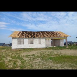 Mistrz-Bud - Budowa domu jednorodzinnego w stanie surowym otwartym: białe bloczki, drewniana konstrukcja dachu, widok z zewnątrz na tle błękitnego nieba i zieleni.